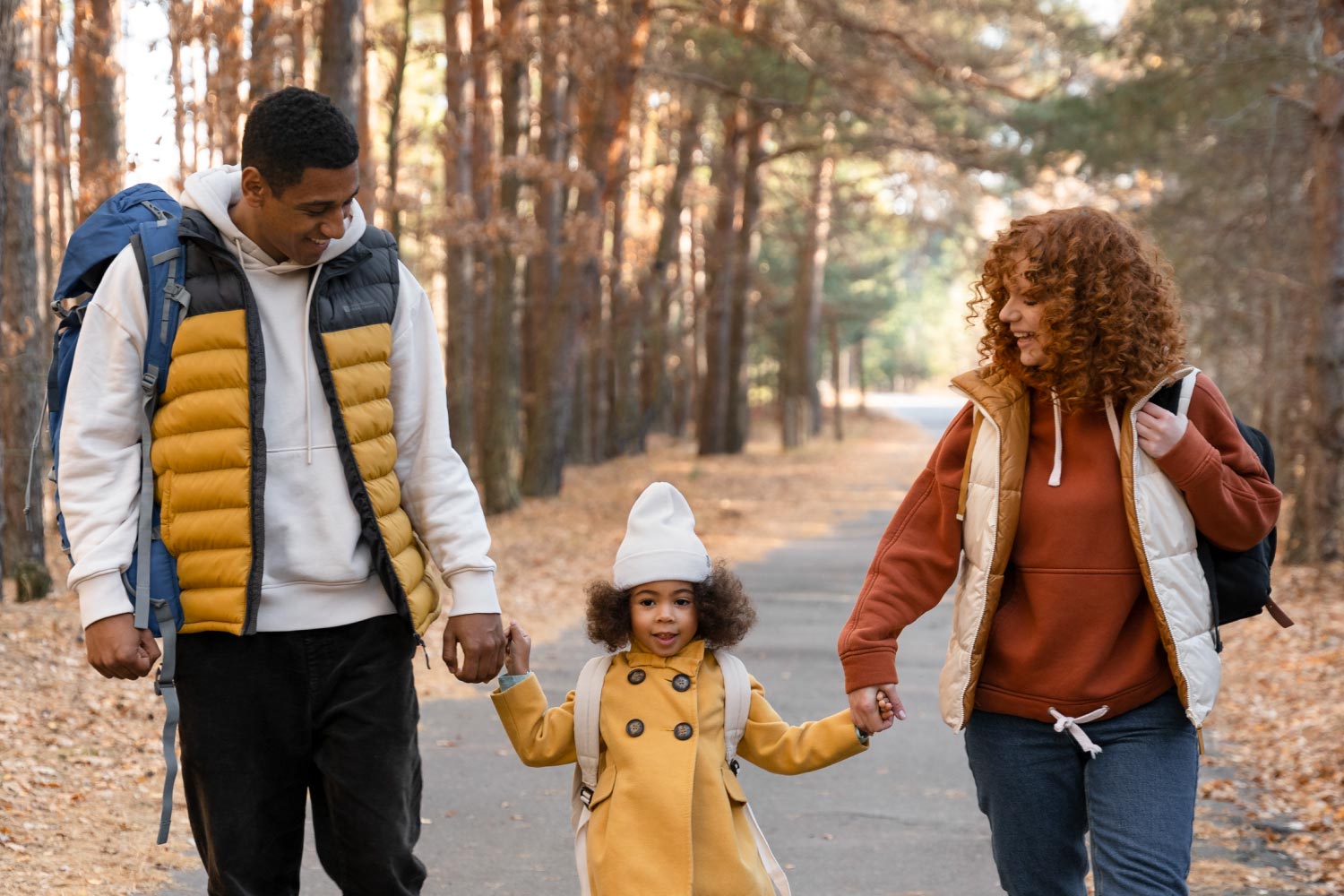 family walks benefit everyone in many holistic ways. A family of 3 walks in a wooded area holding the hand of their young daughter after school.