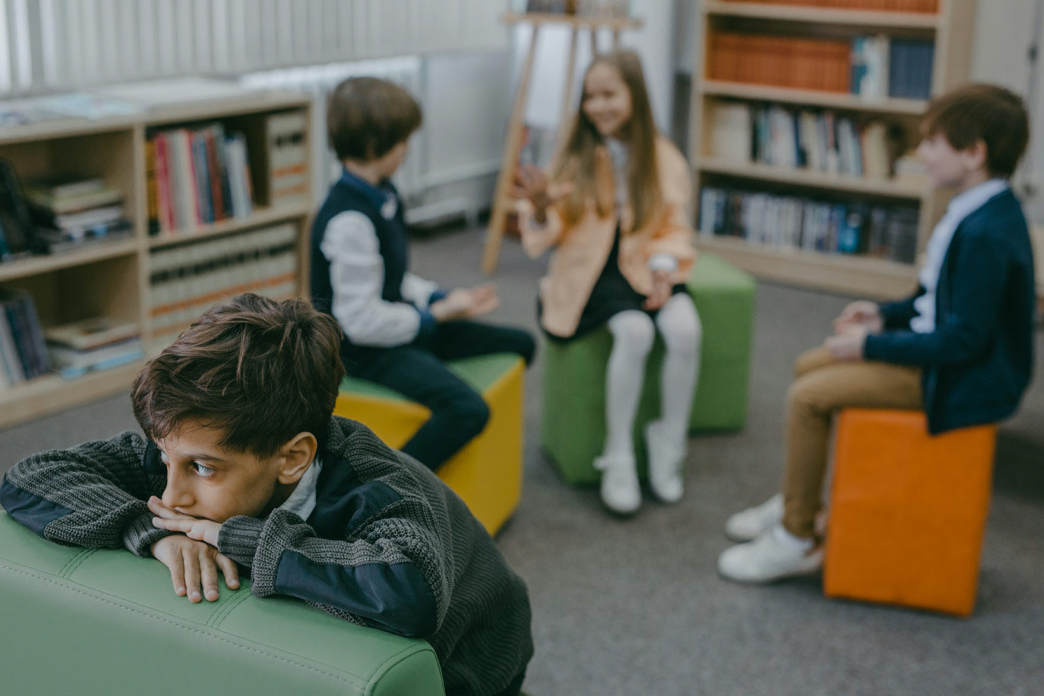 Sensory Overload vs. Behavioral Problems. A boy sits by himself in a library with friends in the background.