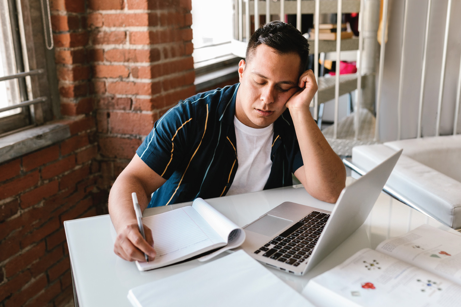 College student sitting in a loft apartment with books and a laptop open visibly stressed