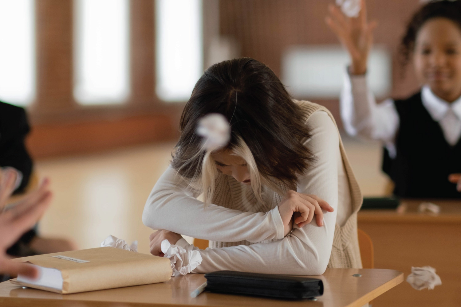 A young teen sits at a lunch table alone while other teens bully her by throwing paper balls at her